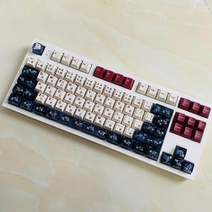 May include: White mechanical keyboard with a variety of keycaps. The keycaps are white, cream, burgundy, and navy blue. Some keycaps have Japanese characters. The keyboard has a white frame.