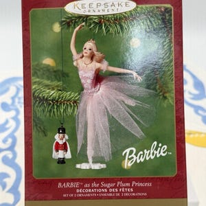 May include: A Hallmark Keepsake Ornament box featuring Barbie as the Sugar Plum Princess and a small nutcracker ornament. The packaging displays a blonde doll in a pink tulle ballet dress and a miniature nutcracker figure with a white cape.
