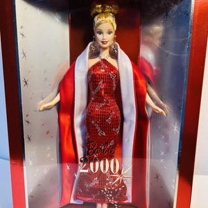 May include: A Barbie Collector Edition doll from the year 2000, presented in a red box. The doll wears a red sequined gown and a white-trimmed red shawl. The box features the text "Barbie 2000" and "Collector Edition".