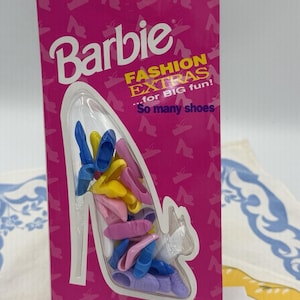 Puede incluir: Un set de Barbie Fashion Extras en su empaque original con una colección de zapatos de muñeca en miniatura de plástico en azul, amarillo, rosa, morado y blanco. El cartón rosa muestra el logo de Barbie y el texto Fashion Extras y So many shoes.