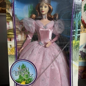 Puede incluir: Una muñeca Barbie Glinda la Bruja Buena con un vestido de satén rosa, mangas abullonadas transparentes y una sobrecapa de falda plateada brillante. La muñeca lleva una corona plateada alta y una gargantilla en forma de estrella.