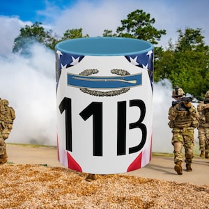 May include: White ceramic mug with a blue interior, featuring a design with the number "11B" in bold black letters. The mug has an American flag design and a military emblem. Soldiers are in the background.