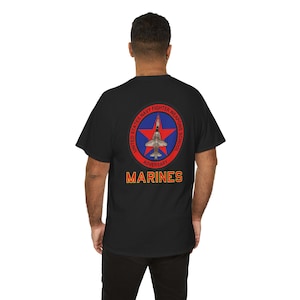 Puede incluir: Camiseta negra con un emblema circular en la espalda. El emblema presenta una estrella roja, un avión a reacción y el texto "United States Navy Fighter Weapons School Adversary Marines" en blanco y naranja. La camiseta tiene cuello redondo.