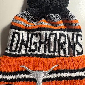 May include: Orange and black knit beanie with the word "LONGHORNS" in large black letters. The beanie features a white embroidered longhorn logo and a black pom-pom on top. The hat has orange, black, and white stripes.