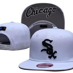 May include: A white snapback cap featuring a black embroidered Chicago White Sox logo on the front, a black New Era flag on the side, and a grey under-brim with the word Chicago in black script. The back includes an MLB logo patch and snap closure.