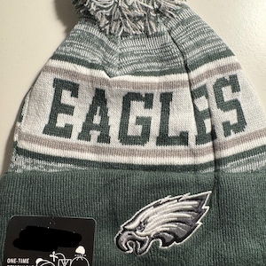 Könnte beinhalten: Eine grüne, graue und weiße Strickmütze mit dem Wort "EAGLES" in großen Buchstaben. Die Mütze hat einen Bommel oben und ein gesticktes Adlerlogo. Ein Etikett ist an der Mütze befestigt.