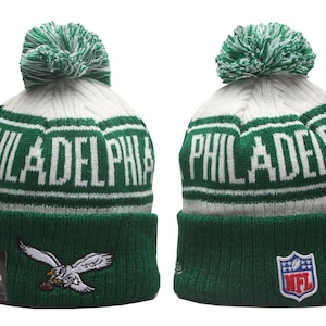 Könnte beinhalten: Eine grün-weiße Strickmütze mit Bommel, dem Wort PHILADELPHIA in weißen Buchstaben, einem gestickten Adler-Logo auf dem vorderen Umschlag und einem NFL-Schild-Aufnäher am seitlichen Umschlag, die Vorder- und Seitenansicht zeigend.