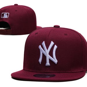 May include: A maroon baseball cap featuring a white embroidered NY logo on the front, a white MLB logo on the back, and a snapback closure. The cap has a structured crown and a flat brim, with a small white logo detail on the side panel.