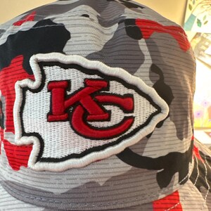 May include: A camouflage-patterned baseball cap with a white and red embroidered logo. The logo features the letters "KC" in red, inside a white arrowhead. The cap's pattern includes shades of gray, black, and red.