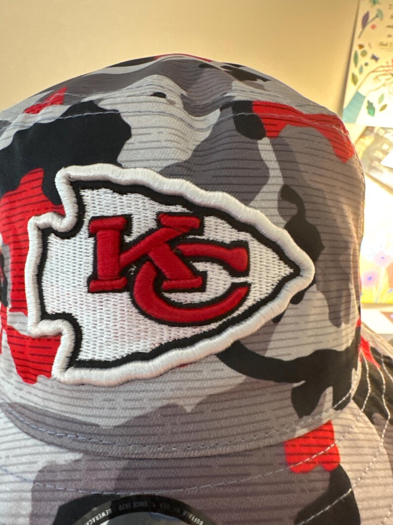 May include: A camouflage-patterned baseball cap with a white and red embroidered logo. The logo features the letters "KC" in red, inside a white arrowhead. The cap's pattern includes shades of gray, black, and red.