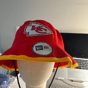 Op de afbeelding: Rode vissershoed met een gele rand en een zwart trekkoord. De hoed heeft het logo van de Kansas City Chiefs en een New Era-patch. De hoed wordt getoond op een mannequinhoofd.