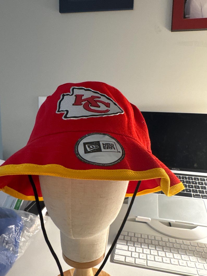 Op de afbeelding: Rode vissershoed met een gele rand en een zwart trekkoord. De hoed heeft het logo van de Kansas City Chiefs en een New Era-patch. De hoed wordt getoond op een mannequinhoofd.