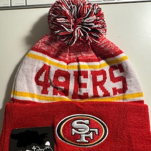 Peut inclure: Un bonnet en tricot rouge, blanc et jaune avec le texte "49ERS" en grandes lettres blanches. Le bonnet présente un écusson du logo de l'équipe et un pompon multicolore. Une étiquette avec du texte est attachée.