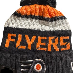 Peut inclure: Un bonnet en tricot noir, orange et blanc, avec le mot "FLYERS" en lettres orange. Le bonnet a un pompon noir et le logo des Flyers de Philadelphie.