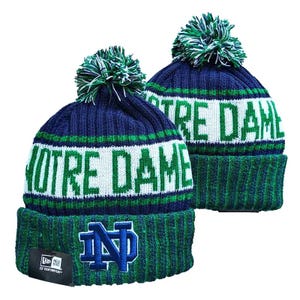 Op de afbeelding: Twee marineblauwe en groene gebreide mutsen met pompons. De mutsen hebben een witte band met de tekst NOTRE DAME in groene letters. De omgeslagen boord toont een blauw geborduurd ND-logo en een zwart New Era-label aan de voorzijde.