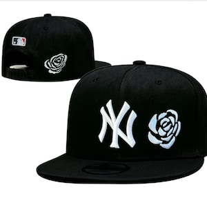Könnte beinhalten: Eine schwarze Baseballkappe mit einem weiß gestickten New York Yankees Logo und einer weißen Rosengrafik auf der Vorderseite. Die Kappe hat einen flachen Schirm, einen Snapback-Verschluss und ein MLB-Logo-Patch auf der Rückseite mit einer Rose.
