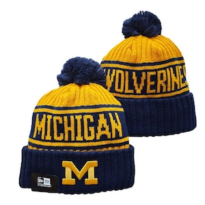 May include: Two navy blue and yellow knit beanies with pom-poms. One beanie features the word MICHIGAN in yellow text and a yellow M logo on the cuff. The other beanie displays the word WOLVERINES in yellow text. Both have a ribbed cuff design.