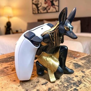 May include: A black and gold Anubis statue holding a white and black game controller. The statue is kneeling with its head and ears upright. The controller is white with black buttons. The statue is on a wooden table.