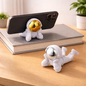May include: White astronaut-shaped phone holder with a gold-colored helmet supporting a black smartphone. A second astronaut figure with a silver helmet rests nearby. The figures are on a wooden surface.