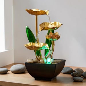 May include: An indoor water fountain with a tiered design. The fountain has gold-coloured lotus-shaped bowls and a black square base. Water flows from the top, creating a soothing ambiance. Green leaves and small stones add to the natural aesthetic.
