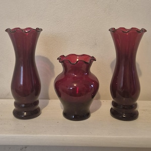 Könnte beinhalten: Ein Set aus drei dunkelroten Glasvasen. Zwei Vasen sind hoch mit schlanken Hälsen und ausgestellten, gewellten Rändern. Die mittlere Vase ist kürzer und breiter mit bauchigem Körper und passendem gewelltem Rand. Alle drei haben eine glatte Oberfläche.