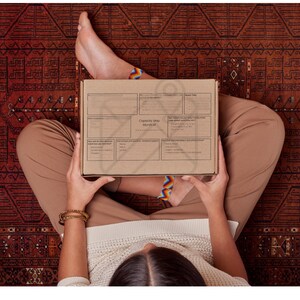 May include: A person holding a cardboard box with printed text, sitting on a patterned rug. The box is tan with a diagram and text. The person is wearing tan pants and a cream-colored sweater. Rainbow-colored bracelets are visible on the ankles.