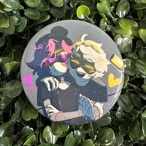 May include: A circular pin-back button featuring a stylized illustration of two characters embracing. One character has dark hair with pink highlights and glowing pink eyes, while the other has blonde hair and glowing yellow eyes. Pink and yellow hearts surround them.