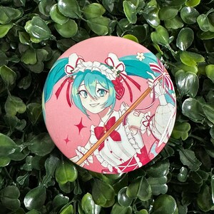 May include: A circular pinback button featuring a colorful anime-style illustration of a character with teal twin-tails, wearing a white and red maid-inspired outfit with a ruffled headpiece, set against a solid pink background with star accents.