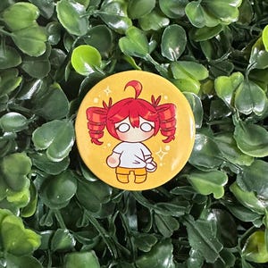 May include: A circular pinback button featuring a yellow background and a cartoon illustration of a character with red hair styled in twin tails, wearing a white shirt and yellow shorts, accented by small white star-like sparkles around the figure.