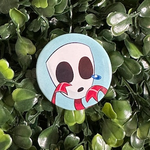 Pode incluir: Um broche redondo com fundo azul-claro apresentando uma personagem de desenho animado com máscara branca, grandes olhos ovais escuros e uma lágrima azul. A personagem tem detalhes vermelhos e uma boca pequena, em estilo gráfico plano.
