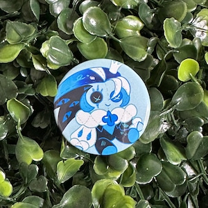 May include: A circular pinback button featuring a stylized illustration of a blue-toned character with a crown, stitched eye details, and a dark cape with multiple eye motifs, set against a light blue background with white accents.