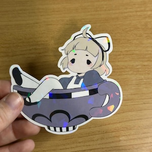 Puede incluir: Una pegatina con un personaje de dibujos animados en una taza de té. El personaje tiene pelo rubio, un lazo blanco y lleva un vestido azul. La taza de té gris tiene un borde a rayas blanco y negro y un efecto holográfico.