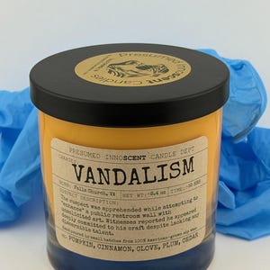 Puede incluir: Una vela en tarro con tapa negra y etiqueta que dice "Vandalismo". La vela es amarilla con base azul oscuro. La etiqueta incluye las notas de fragancia: calabaza, canela, clavo, ciruela y cedro.