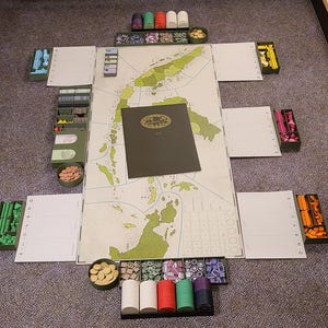 Puede incluir: Configuración de un juego de mesa con un tablero de juego estilo mapa, varias piezas de juego de colores y tableros de jugador. El tablero está rodeado de bandejas que contienen componentes del juego. Un libro verde oscuro está en el centro.