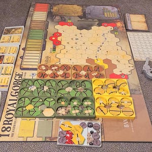 Può includere: Un set di giochi da tavolo, con una grande tavola da gioco con una griglia esagonale, vari pezzi di gioco, carte e vassoi portaoggetti. Il tabellone ha un design a mappa con toni rossi e marroni. Il gioco si intitola "18RoyalGorge".
