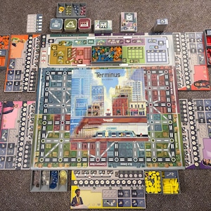 Puede incluir: Juego de mesa "Terminus" con un diseño de ciudad. El tablero presenta un paisaje urbano detallado con carreteras, edificios y un tren. Varios componentes del juego, como cartas y fichas, se organizan alrededor del tablero.