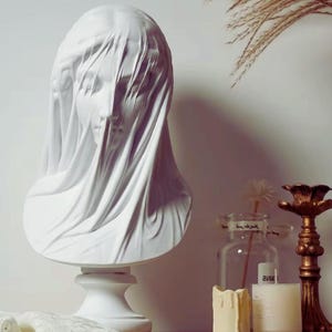 May include: A white bust sculpture of a veiled figure. The draped fabric covers the head and shoulders, creating a sense of mystery. The sculpture sits on a white pedestal. Decorative candles and a candlestick are in the background.