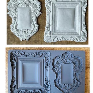May include: Two white, ornate picture frames with detailed scrollwork and floral accents sit on a wooden surface. Below, a grey silicone mould features two more frames with similar designs. The frames are rectangular with a central recessed area.