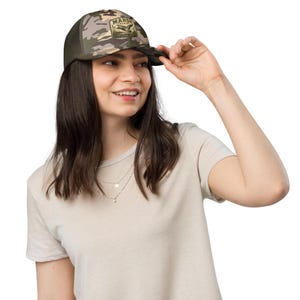 Può includere: Un cappellino da baseball mimetico con retro in rete verde oliva scuro e pannello frontale strutturato. La parte anteriore presenta una toppa rettangolare con il testo MARSH COUNTRY e l'illustrazione di un'anatra che vola sopra dei canneti.