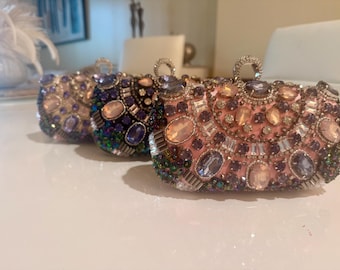 May include: Three embellished evening clutches in a row. The bags are covered in sparkling gemstones in shades of pink, purple, and iridescent green. Each clutch has a small, jeweled handle.