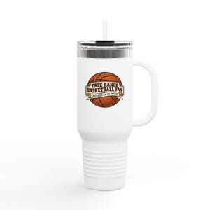 May include: White travel mug with a handle and a straw. The mug has a basketball graphic with the text "FREE RANGE BASKETBALL FAN" and "JUST HERE FOR THE SNACKS". The mug is designed to keep drinks hot or cold.