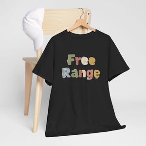 Puede incluir: Una camiseta negra con las palabras "Free Range" en letras coloridas de estilo patchwork. La camiseta está colgada en una percha de madera, con una gorra de béisbol blanca apoyada en una silla de madera en el fondo.