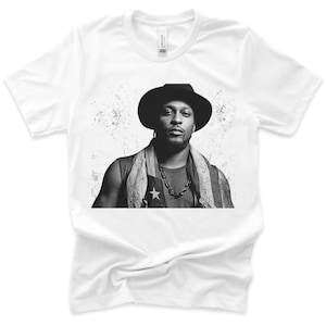 Può includere: T-shirt bianco con un ritratto in bianco e nero di una persona con cappello, catena e sciarpa fantasia. Il ritratto ha uno sfondo in stile grunge. La maglietta è realizzata in un materiale morbido.