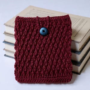 May include: A burgundy crochet book cover with a textured pattern and a blue button closure. The cover is shown resting on a stack of books, showcasing its handmade quality and protective design for books.