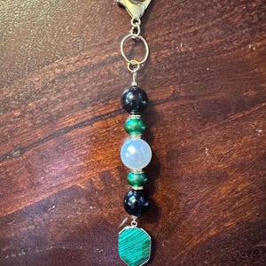 May include: A beaded keychain with a gold-colored clasp. The keychain features black, green, and white beads, and a green malachite stone pendant. The beads are strung on a silver-colored wire.