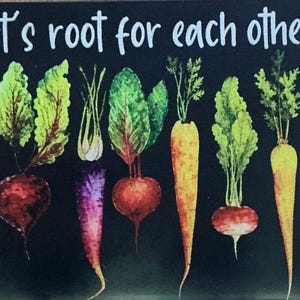 May include: A rectangular sign with a black background featuring the white text Let's root for each other above a row of watercolor-style vegetables including parsnips, red beets, purple carrots, orange carrots, and radishes with green leafy tops.
