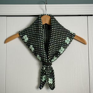 May include: A black and white knit scarf with a repeating pattern and light green floral accents. The scarf is knotted at the bottom and hangs from a wooden hanger.