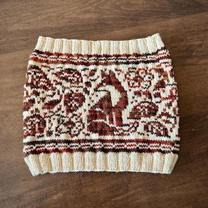 May include: A knitted neck warmer featuring a repeating pattern of foxes and foliage in shades of rust, brown, and cream. The accessory has a ribbed cream-colored border at the top and bottom edges, with a consistent colorwork design throughout the center.