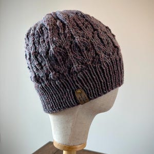 May include: A knitted beanie in shades of gray, burgundy, and purple, displayed on a mannequin head. The hat features a textured cable knit design and a small leather tag with text. The beanie is a winter accessory.