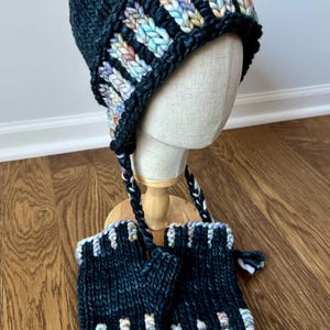 May include: A dark charcoal knit hat with earflaps and braided ties, paired with matching fingerless gloves. Both items feature a thick, chunky texture and a decorative band of variegated yarn in pastel shades of blue, pink, and yellow.